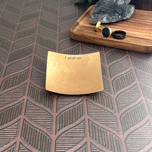bronze square handmade ring dish stamped with "I still do" on the top. Dish is sitting on table with cufflinks and wedding ring in the background
