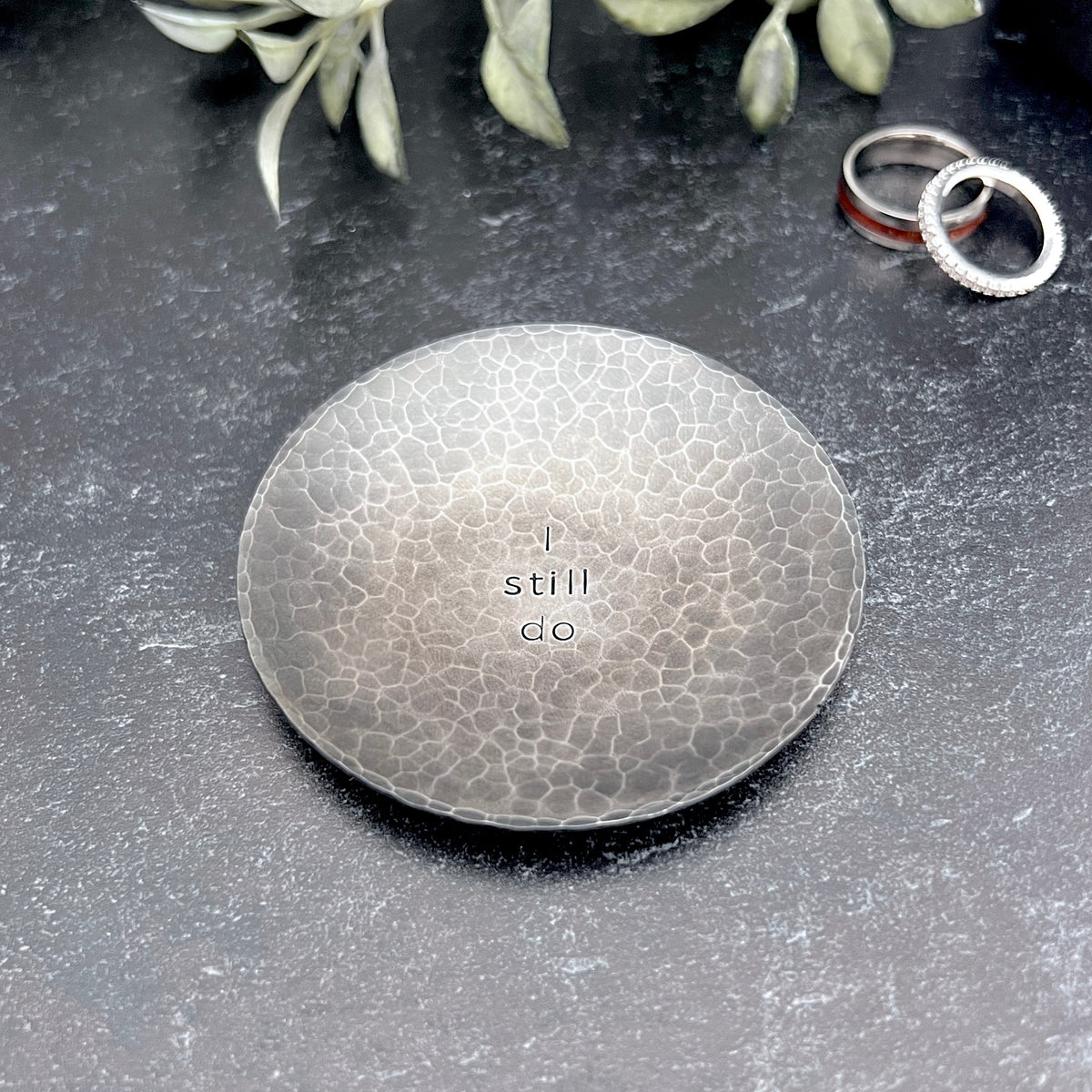 Round hammered blackened steel ring dish stamped with "I still do" in the center. Dish is shown on concrete with wedding rings and driend flowers in the background.