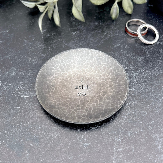 Round hammered blackened steel ring dish stamped with "I still do" in the center. Dish is shown on concrete with wedding rings and driend flowers in the background.