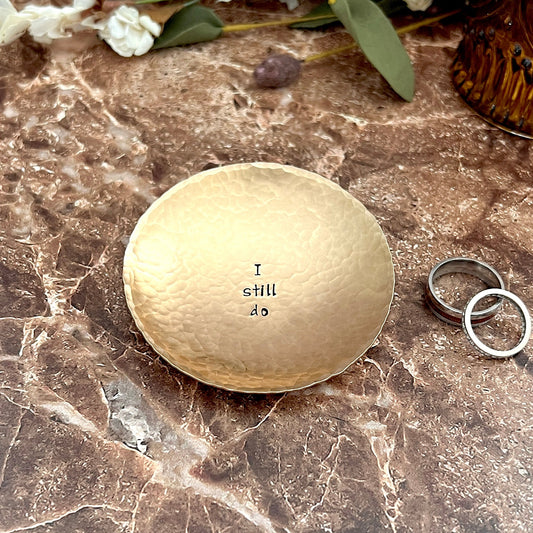 Hammered round bronze ring dish hand-stamped with "I still do" in the center. Dish is shown on marble with wedding rings and dried flowers.