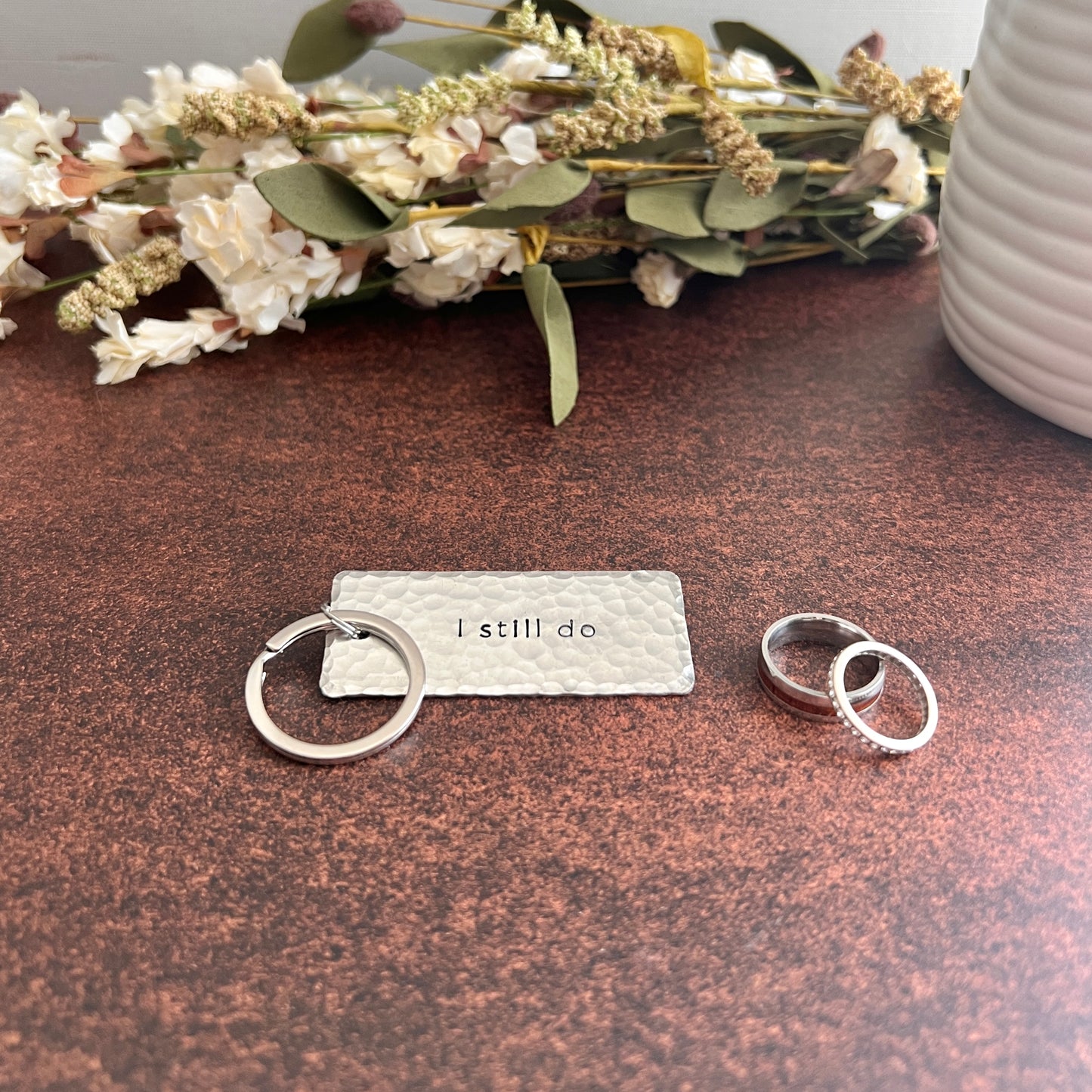 Handcrafted silver-toned "I still do" keychain with hammered texture and silver keyring, styled on a marble surface with wedding rings and dried florals.
