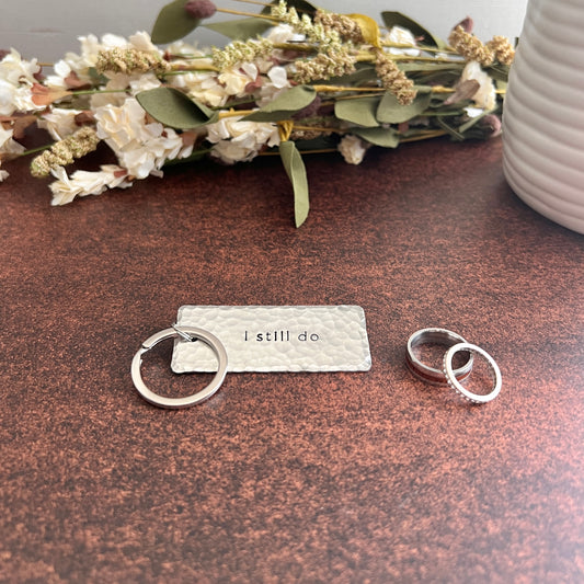 Handcrafted silver-toned "I still do" keychain with hammered texture and silver keyring, styled on a marble surface with wedding rings and dried florals.
