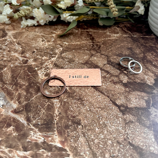 Handcrafted copper "I still do" keychain with hammered texture and copper keyring, styled on a dark surface with wedding rings and dried florals.