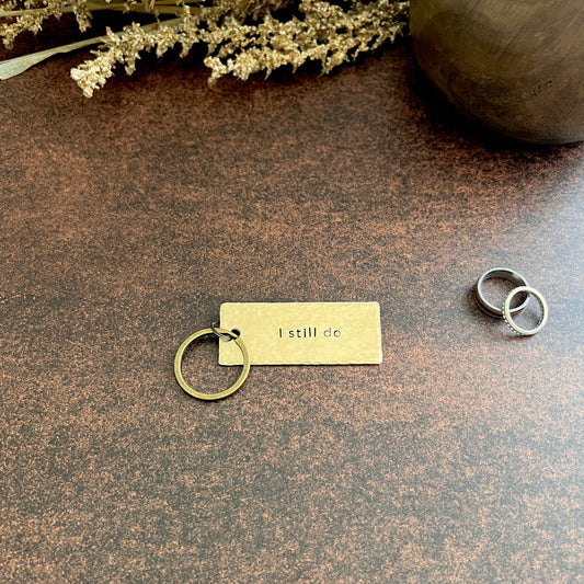 Handcrafted bronze "I still do" keychain with hammered texture and bronze keyring, styled on a dark surface with wedding rings and dried florals.