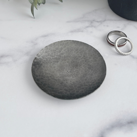 Personalized hammered and darkened steel round ring dish. Dish has 11 in Roman numerals hand-stamped in the center with wedding rings next to the dish.
